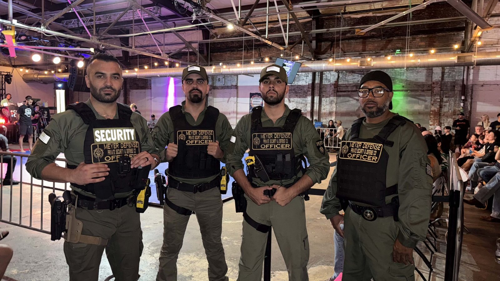 Four security officers from Mayer Security Services standing together in a venue, wearing tactical vests with "SECURITY" and "OFFICER" labels, equipped with radios and sidearms, set against a lively event backdrop.
